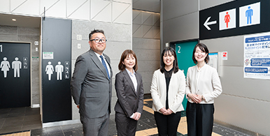 ゆりかもめ　東京ビッグサイト駅の男女共用トイレの前で、座談会のメンバー4人が並んで立ち微笑んでいる