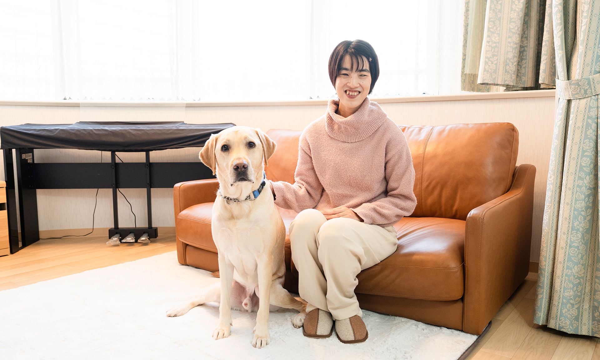 室内でソファに座り微笑む鈴木さんと、傍に座る盲導犬のデイズくん